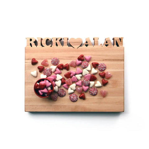 The Handcrafted Wooden Personalized Cutting Board by Words with Boards, made in the USA, features RICKI ♥ ALAN cut out at the top and is shown holding assorted heart-shaped candies—a charming and practical kitchen accessory.