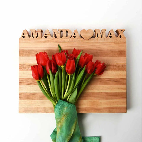 A bouquet of red tulips wrapped in green cloth sits on a Handcrafted Wooden Personalized Cutting Board – Large by Words with Boards, engraved with AMANDA ♥ MAX, creating a charming kitchen display against a white background.