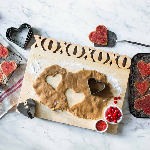 Heart-shaped cookie cutters press into dough on a Handcrafted Wooden Personalized Cutting Board – Large by Words with Boards. Decorated heart cookies, red sprinkles, and candies surround the kitchen scene on a marble countertop.