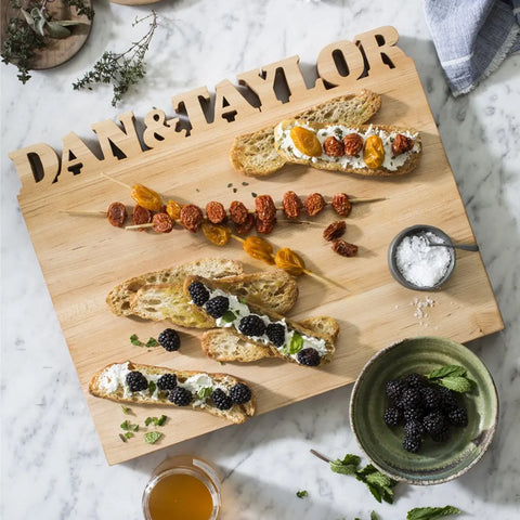 A Handcrafted Wooden Personalized Cutting Board – Large by Words with Boards displays assorted toasts, berries, cheese, herbs, skewered tomatoes, dried fruits, salt, honey, and blackberries on a marble kitchen counter. Made in the USA.