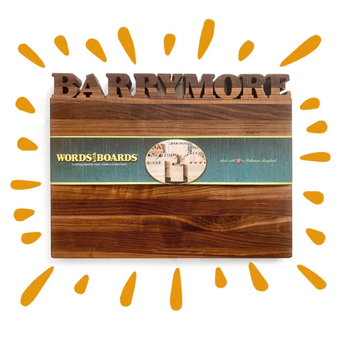 The Handcrafted Wooden Personalized Cutting Board – Large by Words with Boards features “BARRYMORE” in bold 3D letters, a green branded label, and orange illustrated rays—an ideal kitchen display. Made in the USA from American hardwoods.