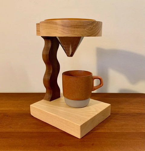 The Squiggle Pour Over by Shop Cat Woodworks features a handmade wavy wood support holding a ceramic dripper above a brown mug, all crafted from reclaimed wood and displayed on a wooden surface against a plain wall.