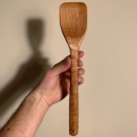A hand holds up The Natural Spatch by Shop Cat Woodworks, a handmade wooden spatula, against a plain beige background as its shadow falls on the wall.