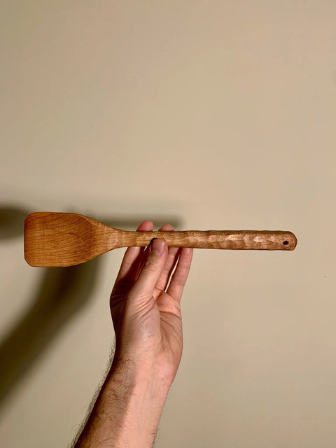 A hand holds The Natural Spatch by Shop Cat Woodworks, a handmade spatula crafted from reclaimed hardwood, with its handle and flat end clearly visible against a plain beige background.