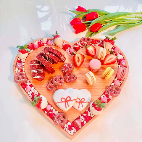 The Handcrafted & Heart Shaped Wooden Cheese Board by Words with Boards, topped with cookies, chocolate-covered strawberries, macarons, candies, and heart-shaped treats. Red tulips rest beside the charcuterie board on a white surface.