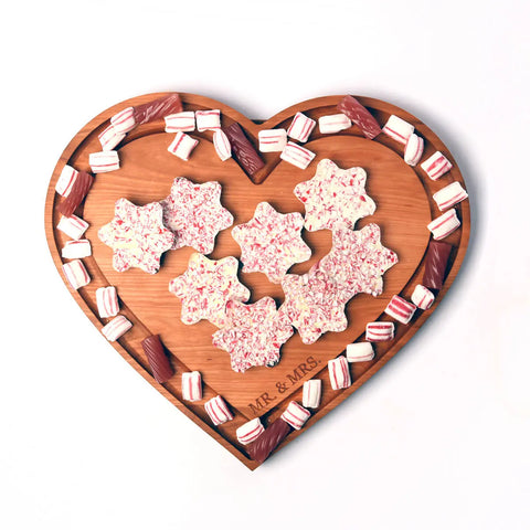 The Handcrafted & Heart Shaped Wooden Cheese Board by Words with Boards displays peppermint star-shaped treats at its center, surrounded by striped peppermint candies and chocolate pieces along the edges. Made in the USA.
