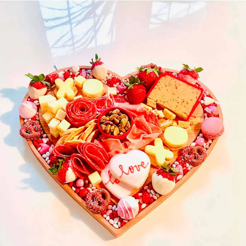 The Words with Boards Handcrafted Heart Shaped Wooden Cheese Board displays cheeses, meats, strawberries, nuts, pretzels, candies, and an I love cookie arranged festively on a white surface. Made in the USA.