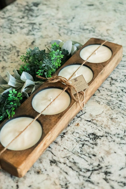 The Handmade 4-Hole Dough Bowl Candle by Shanti + Tula, featuring 100% soy wax in a rustic cheese mold style, is displayed on a speckled granite countertop with green artificial plants. Made in the USA.