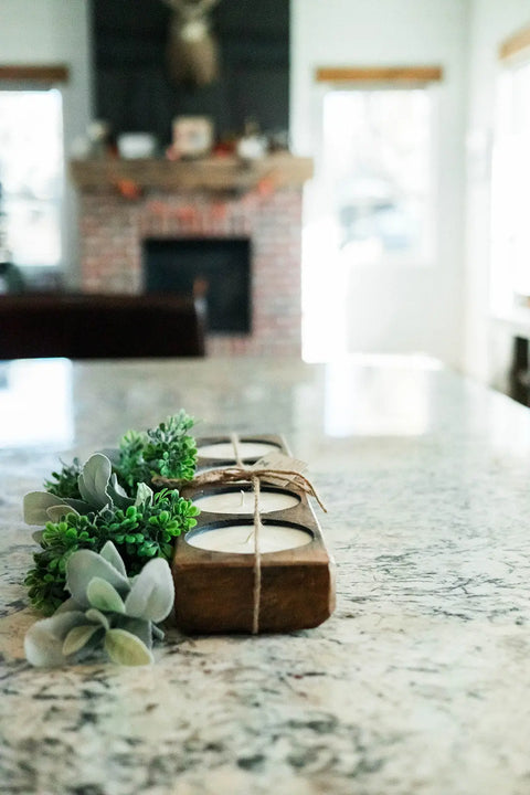 A Handmade 4-Hole Dough Bowl Candle by Shanti + Tula, crafted in rustic cheese mold style with 100% soy wax, is displayed on a marble countertop in a bright living room with a brick fireplace in the background.