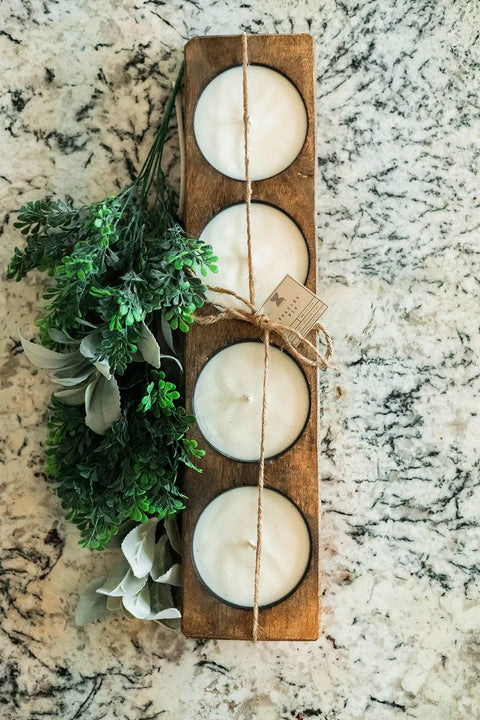 A Handmade 4-Hole Dough Bowl Candle by Shanti + Tula, crafted in a rustic cheese mold style from 100% soy wax, sits on a marbled surface tied with twine, accompanied by green and white artificial foliage. Made in the USA.