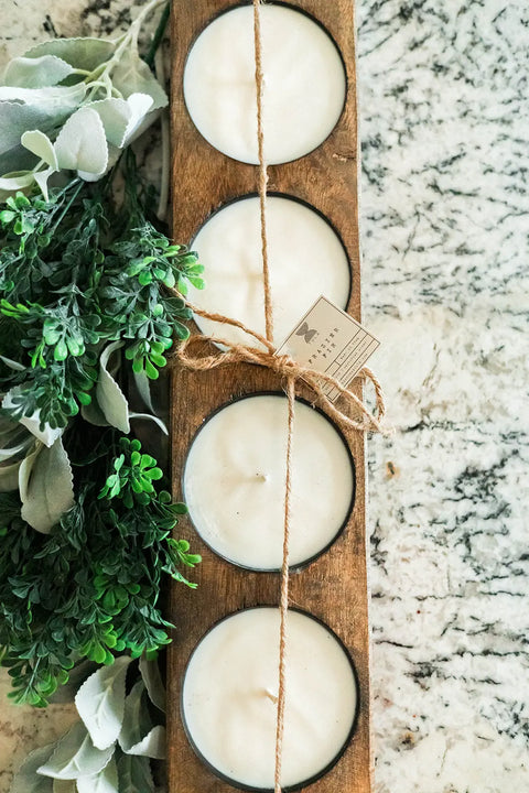 The Handmade 4-Hole Dough Bowl Candle by Shanti + Tula features a rustic cheese mold style and holds four round white 100% soy wax candles, tied with twine and displayed on marble beside green and silver faux foliage.