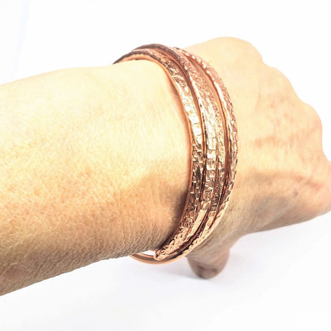 A hand displays the 2 Piece Handmade Interlocking Full Overlap Copper Bangle Set by Alexa Martha Designs, featuring textured, shiny hammered copper bangles against a white background.