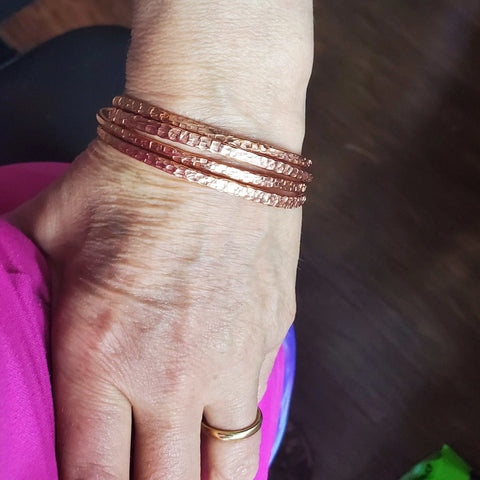 A hand wearing a gold wedding band and the 2 Piece Handmade Interlocking Full Overlap Copper Bangle Set by Alexa Martha Designs rests on pink fabric, set against a dark, blurred background.