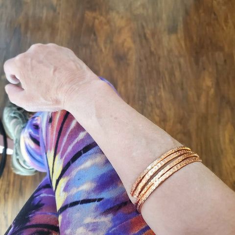 A person wearing colorful tie-dye leggings and the 2 Piece Handmade Interlocking Full Overlap Copper Bangle Set by Alexa Martha Designs sits cross-legged on a wooden floor.