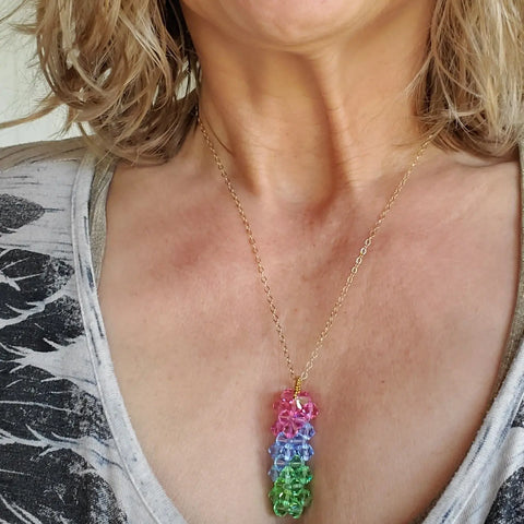 A woman wears the Alexa Martha Designs Super Sparkly Crystal Pillar Pendant in green, blue, and rose with an optional necklace, shown from shoulders to chest over a patterned shirt.