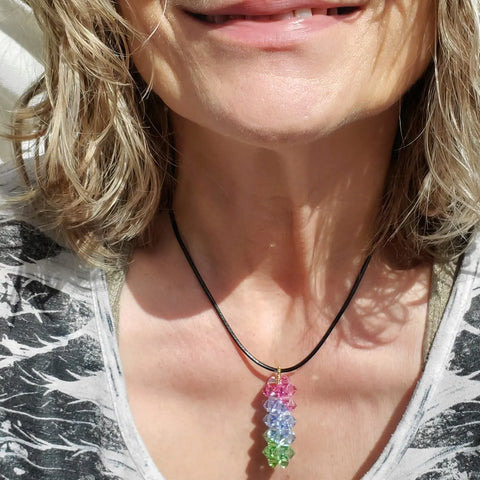 A woman with wavy blonde hair smiles in sunlight, wearing the Alexa Martha Designs Super Sparkly Crystal Pillar Pendant in Green Blue and Rose on a black cord. Only her mouth, neck, and upper chest are visible.