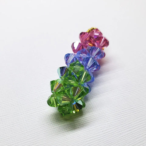 A Super Sparkly Crystal Pillar Pendant in green, blue, and rose by Alexa Martha Designs is displayed vertically on a white textured background, resembling faceted flower shapes. Optional necklace available.