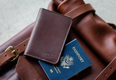 The Urban Southern Passport Wallet, a U.S. passport, and a brown leather bag with a gold buckle sit on a light surface. The wallet partly covers the passport—ideal travel essentials for style and convenience.