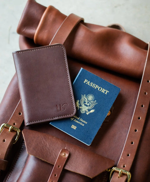 A Passport Wallet by Urban Southern sits atop a brown full-grain leather backpack with a U.S. passport, displaying travel essentials perfectly arranged for your next adventure.