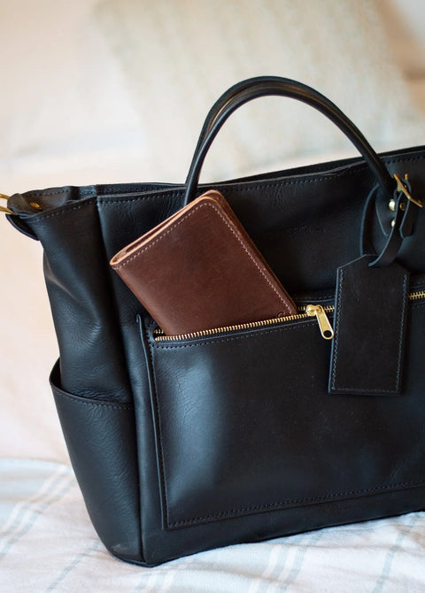 An Urban Southern Passport Wallet peeks from the front zippered pocket of a black full-grain leather handbag, resting on a light blanket—the perfect travel companion.