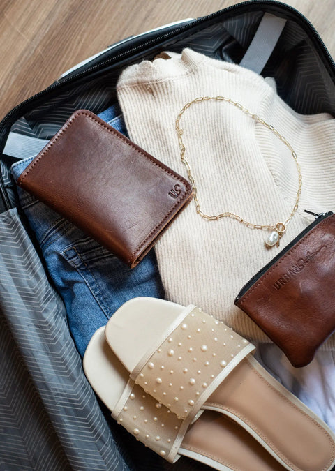 Open suitcase with a folded cream sweater, blue jeans, pearl-studded sandals, gold chain necklace with pearl, and Urban Southerns Passport Wallet and matching coin pouch—your ideal travel companion.
