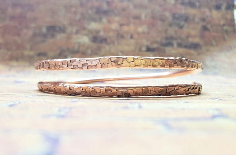 The Full Overlap Copper Bangle by Alexa Martha Designs features two thin, handmade hammered copper bangles with a textured surface, shown against a blurred earth-toned background.