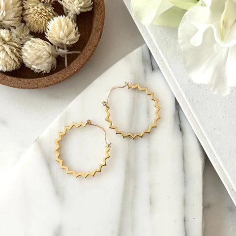 A pair of Sarah Cecelia GOLD POINTE HOOPS, 14K gold plated earrings, displayed on white marble next to dried flowers and a white petal.