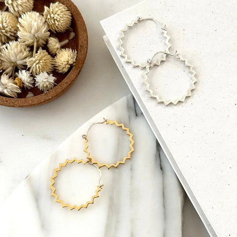 Two pairs of zigzag hoop earrings, including Sarah Cecelia’s GOLD POINTE HOOPS, are displayed on a white marble surface and textured board, with a small bowl of dried flowers in the upper left corner.