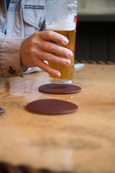 A person’s hand holds a tall glass of beer above a Sebago Camp Coaster by Rogue Industries, made in Maine, on a wooden table with another coaster nearby.