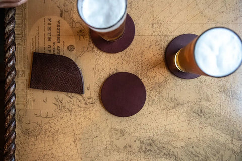 Two tall beers rest on Rogue Industries Sebago Camp Coasters, crafted from top grain leather, atop a vintage nautical map table with a brown leather wallet nearby.