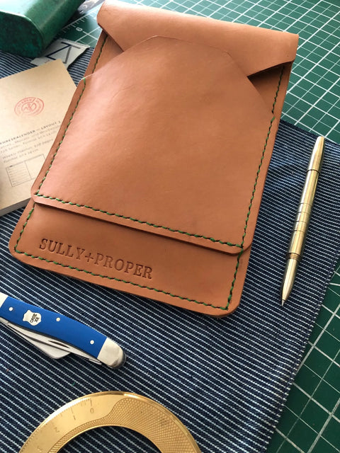 The Sully + Proper Supply Natural Veg Tan Utility Pouch, featuring green stitching and American-made leather, is shown with a gold pen, blue pocket knife, paper card, and brass ruler on a blue striped cloth atop a green cutting mat.