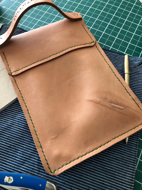 The Sully + Proper Supply Natural Veg Tan Utility Pouch, handmade in Texas with green stitching and a flap, rests on striped fabric. A gold pen and blue pocketknife are nearby, with a green cutting mat in the background.