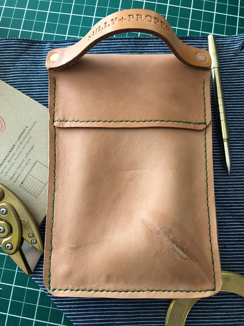 A Sully + Proper Supply Natural Veg Tan Utility Pouch with green stitching and stamped handle rests on striped fabric, alongside a metal pen, brown envelope, brass tool, and curved ruler.