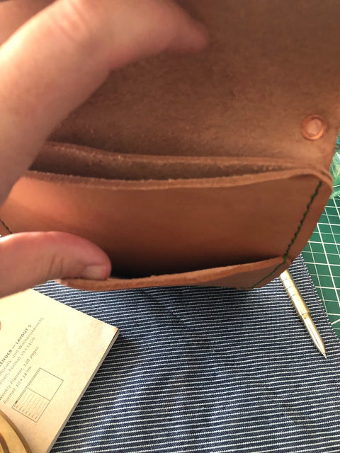 A hand holds open a Sully + Proper Supply Natural Veg Tan Utility Pouch, showing its two compartments. Below, a pen and notebook rest on striped fabric, with a green cutting mat visible in the background.