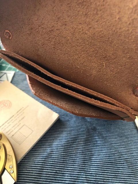 A Sully + Proper Supply Natural Veg Tan Utility Pouch, handmade in Texas, sits open on striped blue fabric, showing its inner pocket. Nearby, a brass drafting tool and a beige envelope or booklet are partially visible.