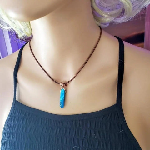 A mannequin showcases Alexa Martha Designs’ Wire Wrapped Pointed Turquoise Agate Leather Necklace for Him and Her, the vibrant pendant standing out against a softly blurred purple and pink backdrop.