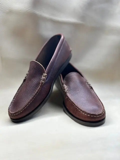 A pair of BBH Loafer shoes in brown pebble grain leather with visible stitching are displayed on a light surface—one upright, the other leaning—to highlight their classic handsewn slip-on style.
