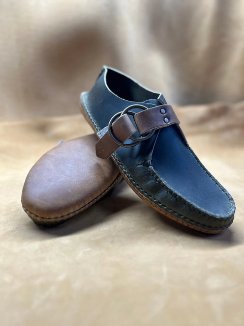 The BBH Mica Mocc is a handmade leather sandal made from premium Horween Chrompak, featuring dark blue uppers, brown straps with buckles, and brown soles. Shown on a tan background with one upright sandal and one tipped on its side.
