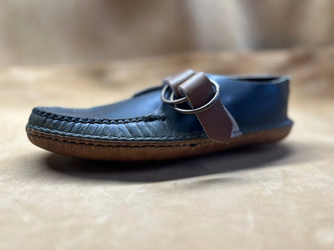 A close-up of the BBH Mica Mocc shows its premium Horween Chrompak leather upper in dark blue, brown sole, and a decorative strap with two metal rings, displayed on a soft beige surface.
