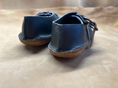 A close-up of the BBH Mica Mocc baby shoes, featuring handmade premium leather moccasin uppers with tan stitching and soft brown soles, displayed on a light brown surface.