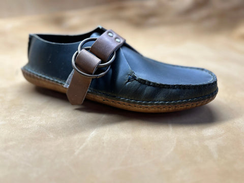 The BBH Mica Mocc is a black premium leather moccasin with a brown sole and a wide brown strap with a silver buckle, shown on a soft tan surface.