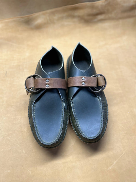 The BBH Mica Mocc is a pair of handmade black premium leather moccasins with brown leather straps and metal buckles, shown on a light brown surface.