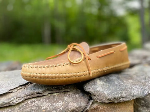 The BBH Twin Sole Old Town Mocc, crafted from tan leather with handsewn detail, sits on a stone surface outdoors against a softly blurred green backdrop of trees and grass.