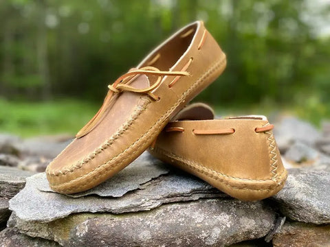 The BBH Twin Sole Old Town Mocc features handsewn tan leather with visible stitching and laces, displayed on stacked stones against a green backdrop—perfect rustic footwear for any setting.