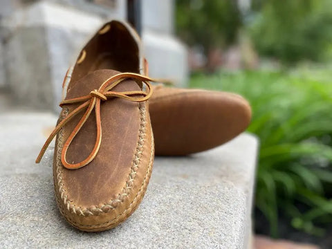 A close-up of the BBH Twin Sole Old Town Mocc—a brown, USA-made leather moccasin with a leather lace bow—rests on an outdoor stone step with blurred greenery in the background.