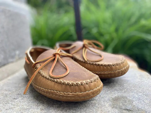 A close-up shows BBH’s Twin Sole Old Town Mocc—USA-made leather moccasins with tan laces—resting on stone outdoors, highlighting rustic style with greenery softly blurred in the background.