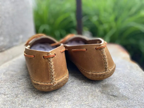 A pair of BBH Twin Sole Old Town Mocc small brown leather moccasins with visible stitching rests on a stone surface, highlighting handsewn craftsmanship against a softly blurred green backdrop.