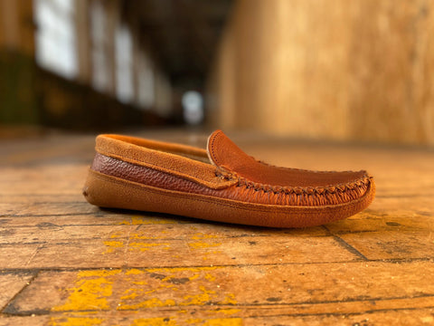 The BBH Northeaster’ is a brown handsewn moccasin slipper made from premium leathers, shown on a wooden floor with blurred walls and windows behind it for a cozy, rustic vibe.
