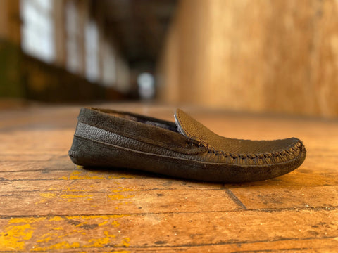 A single “Northeaster’” black handsewn moccasin slipper from BBH rests on a worn wooden floor, its fine stitching and premium leather in sharp focus—a tribute to American craftsmanship against a softly blurred backdrop.