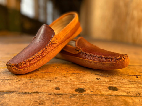 The BBH Northeaster’ brown handsewn moccasin slippers, featuring visible stitching, rest on a rustic wooden surface and display quality American craftsmanship amid warm, soft lighting with a blurred background.